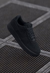Sapatilha Nike Air Force 1 preta com parte superior texturizada, biqueira redonda, perfurações, atacadores planos e sola em borracha. Flutuando acima de uma grelha metálica.