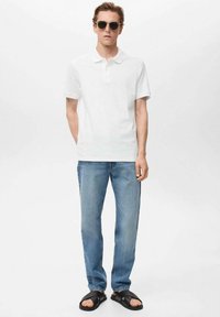 Weißes Poloshirt, hellblaue Jeans und schwarze Sandalen. Das Model trägt dunkle Sonnenbrillen. Einfaches Design, lässige Passform und keine sichtbaren Muster.