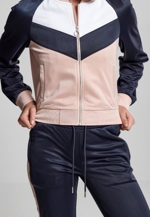 Henkilö, joka pukeutuu väreihin jaetun vetoketjullisen takin, jossa on valkoista, laivastonsinistä ja vaaleanpunaista, ja laivastonsiniset kiristysnauhalla varustetut housut.