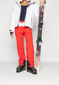 Witte waterdichte jas, marineblauwe top, oranje skibroek met zwarte accenten, zwarte skischoenen, vasthoudend een ski met bindingen.