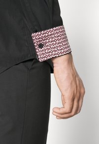 De hand van een man naast een zwarte broek en een zwart overhemd dat opgerold is om een witte en rode patroonvoering met herhaaldelijke "HUGO" tekst te onthullen.