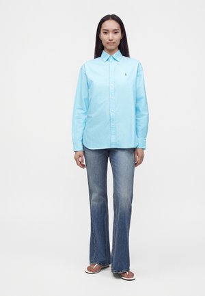 Kvinde står forovervendt iført en lyseblå skjorte med knapper, falmede blå jeans og hvide sandaler på en ensartet hvid baggrund.