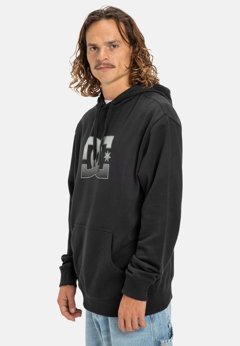 Man with curly hair and a mustache wearing a black hoodie with a DC logo, posed against a plain white background.