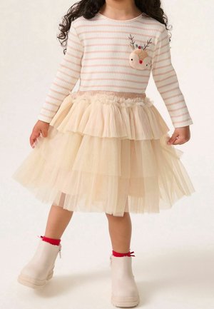 Enfant portant un haut à manches longues crème rayé avec une applique de renne, une jupe en tulle beige superposée, des chaussettes rouges et des bottines crème, debout.