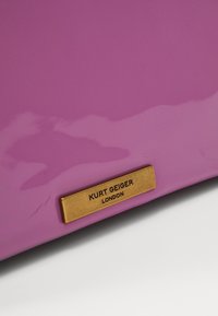 Lila patenterad läderhandväska med en slät yta och en guldmetallplatta med texten "Kurt Geiger London" på framsidan.