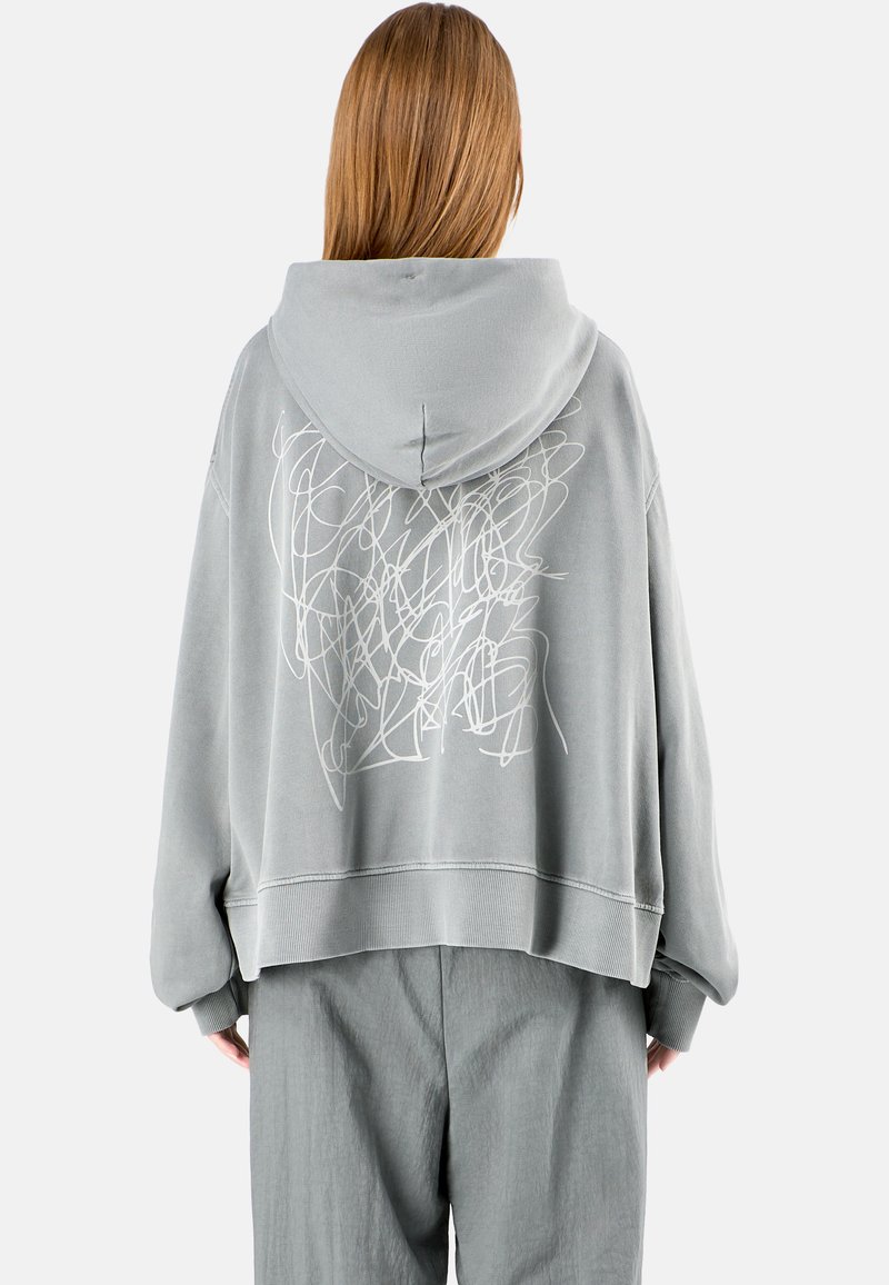 Szary, oversize hoodie z kapturem na sznurku. Posiada abstrakcyjny, biały wzór na plecach oraz ściągacze przy mankietach. Miękka tekstura materiału.