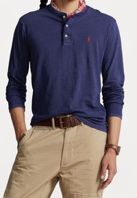 Marinblå långärmad henley-tröja med tre knappar och en röd broderad logotyp; kombinerad med beige byxor och ett brunt läderbälte.