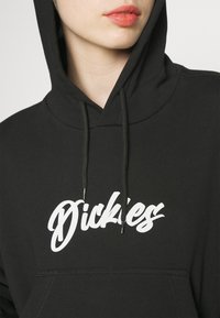 Personne portant un sweat à capuche noir avec un texte blanc "Dickies" sur la poitrine, montrant partiellement le visage et les lèvres avec un rouge à lèvres corail.