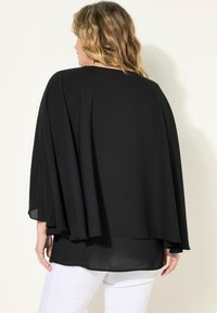 Haut en cape noir en tissu léger, présentant une coupe ample et des manches fluides. Porté sur un pantalon blanc, mettant en avant un design à superpositions.