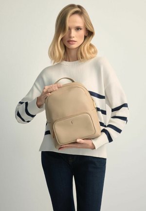 Kobieta o blond włosach, ubrana w biały sweter w czarne paski, trzyma beżowy skórzany plecak na jednolitym jasnym tle.