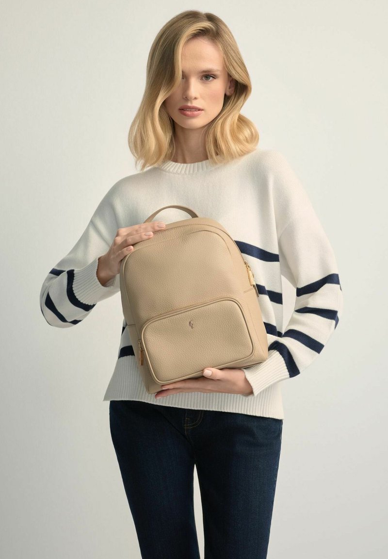 Kobieta o blond włosach, ubrana w biały sweter w czarne paski, trzyma beżowy skórzany plecak na jednolitym jasnym tle.