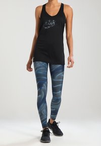 Débardeur noir avec un logo foncé, associé à des leggings bleus à motifs tourbillonnants. Chaussures de sport noires complètent la tenue.