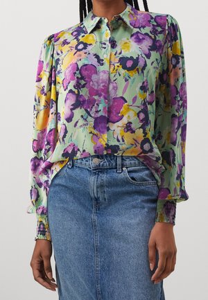 Femme portant un chemisier à manches longues à fleurs aux nuances de violet, jaune et vert, rentrée dans une jupe en denim bleu taille haute.