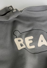 Siva majica z žepom iz mehke tkanine, na sprednji strani velik napis "BEAR" kremne barve in značilni obris medveda. Dva zapirala z zaponkama na ovratniku.