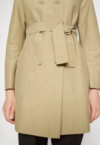 Beżowy trencz z podwójnym zapięciem na guziki, paskiem związanym w talii oraz gładkiego materiału. Widoczne akcenty bocznych szwów i dopasowany krój.