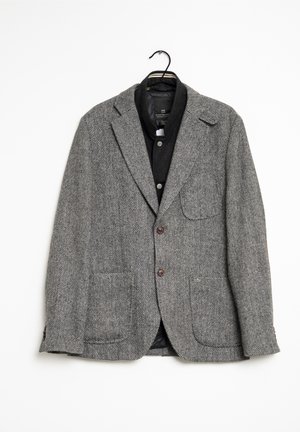 Grauer Fischgrat-Blazer aus strukturiertem Stoff, mit zwei Fronttaschen, braunen Knöpfen und einem geschlitzten Kragen. Hängt an einem schwarzen Haken.