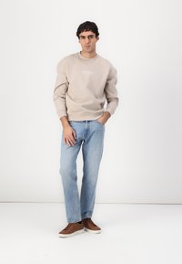 Sweatshirt bej cu manșete și guler cu nervuri, asortat cu blugi drepti de culoare albastru deschis și pantofi sport maro cu tălpi albe.