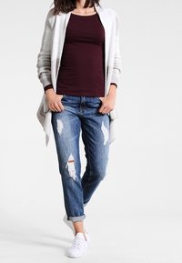 Dlouhý světle šedý kardigan s texturovaným vzorem přes burgundské přiléhavé tričko, spárovaný s roztrhanými modrými džínami a bílými teniskami.