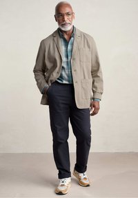 Beige linnen jasje over een teal geruit shirt, gecombineerd met navy broek en multicolor sneakers. Opvallende details zijn een borstzak en de textuur van de stof.