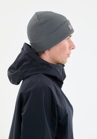Grijze gebreide beanie met een geribbelde textuur en een klein logo-label, gedragen met een marineblauwe hoodie met een trekkoord. Zijprofielweergave.