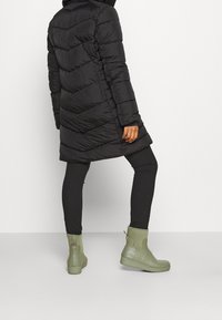 Abrigo de plumas negro con diseño en forma de chevron, hasta la rodilla, combinado con botas verdes impermeables. El conjunto incluye leggings negros ajustados.