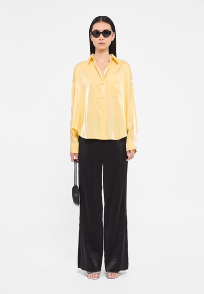 Rumena satenasta oversize srajca z gumbi, črne široke hlače, črne sončne očala, minimalistična črna torbica in srebrni sandal s trakovi.
