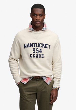 Mężczyzna w beżowej bluzie z napisem „NANTUCKET 954 GRADE”, pod spodem w koszulę w kratę, oliwkowe spodnie i zegarek na rękę, stoi z rękami w kieszeniach.