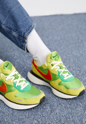 Person iført hvide sokker og opsmøgede blå jeans med grønne, gule og røde Nike-sneakers på blåt tæppe.