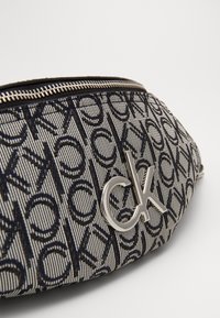 Piccola bustina con zip, caratterizzata da un motivo a logo Calvin Klein in bianco e nero e un emblema "ck" in metallo argentato sulla superficie del tessuto.