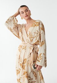 Wikkeljurk in crème met een gouden loofpatroon, lange vloeiende mouwen en een strikceintuur. Heeft een glanzende, ronde gouden clutch in de hand.