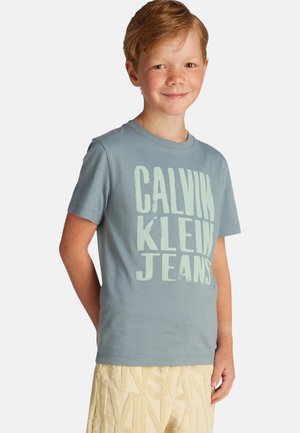 Camiseta de algodón en azul claro con "CALVIN KLEIN JEANS" impreso en grandes letras verdes en contraste. Cuello redondo y mangas cortas.