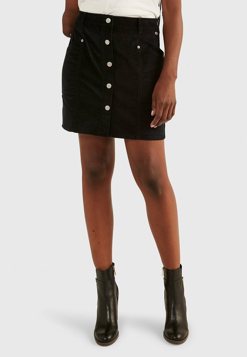 Femme portant une jupe noire boutonnée sur le devant avec des poches et des bottines noires, debout devant un fond uni.