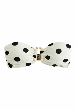 Femme aux longs cheveux foncés portant un bikini bandeau blanc à pois noirs, debout contre un mur beige clair, regardant légèrement vers la gauche.