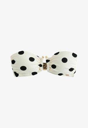 Femeie cu păr lung și închis la culoare, purtând un bikini bandeau alb cu buline negre, stând în picioare lângă un perete bej deschis, privind ușor spre stânga.
