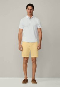 Camisa polo branca, calções amarelos e sapatos castanhos. A polo possui colarinho, enquanto os calções têm um corte reto.