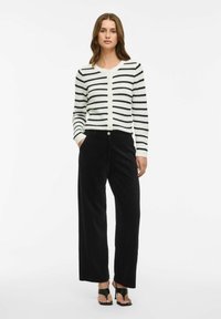 Melna un balta svītraina kardigāna adīta auduma, kombinēta ar plašiem melniem korduroja biksēm. Vienkārša pogu aizdare, minimālistisks dizains.