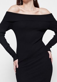 Černé žebrované šaty bez ramínek s dlouhými rukávy, přiléhavý střih a jemná textura. Žádné viditelné vzory ani kovové detaily.