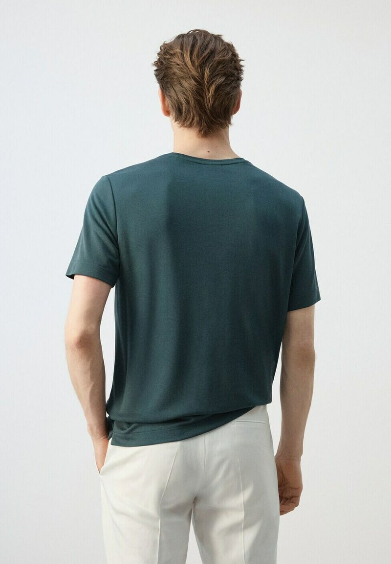 Homme aux cheveux châtain clair portant une chemise à manches courtes vert foncé et un pantalon blanc, vu de dos sur un fond uni.