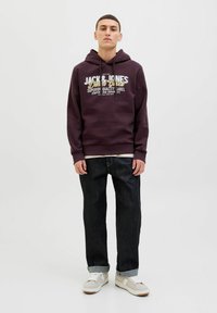 Mørk lilla hættebluse med hvid og gul logo tekst, parret med mørkeblå jeans og lyse sneakers. Hætteblusen har snoretræk og ribbede manchetter.