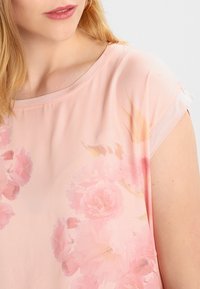 Femme portant un chemisier rose clair avec de doux motifs floraux, des manches courtes et un col rond, sur un fond neutre.
