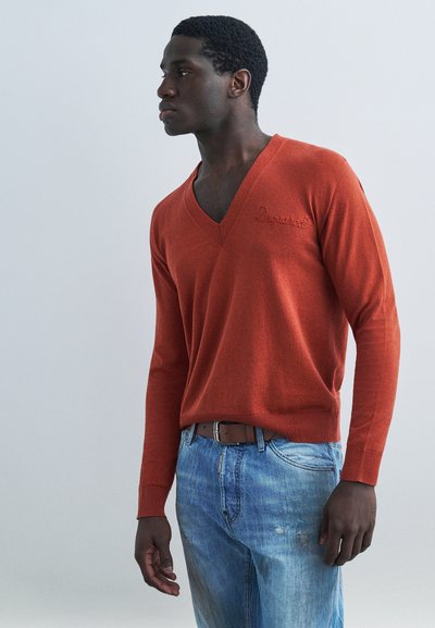 Homme portant un pull à col en V couleur rouille et un jean bleu, debout et regardant vers sa gauche sur un fond clair uni.