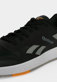 Μαύρο και γκρι αθλητικό παπούτσι Reebok με μαύρα κορδόνια, άσπρη σόλα με πορτοκαλί λεπτομέρεια και λογότυπο Reebok στο πλάι και στη γλώσσα.