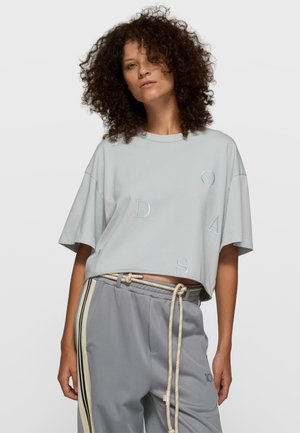 Jasnoszara krótka koszulka z dużymi haftowanymi literami "O", "D", "A", "S". W zestawie z luźnymi szarymi spodniami z bocznymi paskami i sznurkiem.