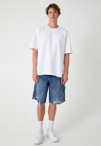 Camiseta blanca de algodón con un corte relajado, combinada con pantalones cortos de mezclilla azul desgastados con bolsillos y dobladillos deshilachados. Lleva puestas zapatillas blancas.
