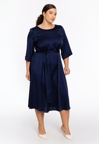 Marineblaue Satinklיד mit rundem Ausschnitt, dreiviertellangen Ärmeln und einem Bindegürtel mit Quasten. Fließende A-Linien-Silhouette und knielanger Saum.