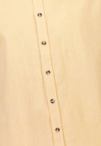 Lichtgele linnen overhemd met zeven metalen knopen aan de voorkant; zachte textuur met een licht kreukelig uiterlijk.