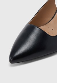 Γυναικείο μαύρο δερμάτινο μπαλαρίνα με μυτερή μύτη, ταμπά διάτρητη επένδυση, σε ανοιχτό φόντο.