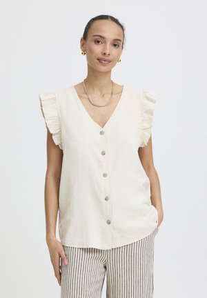 Nő ujjatlan, gombos krémszínű blúzban fodros vállrésszel és csíkos nadrágban, kezét zsebre téve áll.