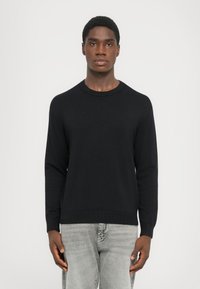 Pulover negru cu guler rotund, confecționat dintr-un material tricotat, cu mâneci lungi și mansete cu model în rilă. Asociat cu blugi gri deschis.