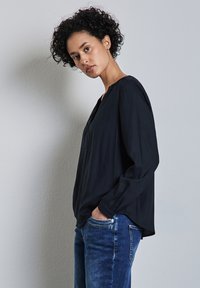 Blusa negra de manga larga con diseño de escote en V y corte holgado, combinada con jeans de mezclilla azules. Textura suave, con detalles mínimos.